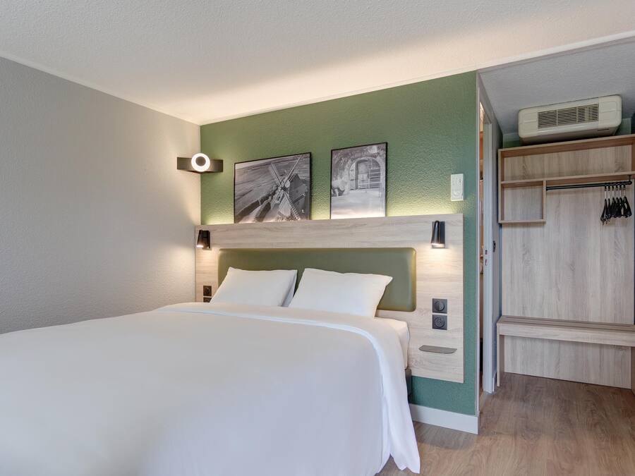 A hotel room with a bed, a wooden headboard, a green accent wall, a mirror, and a wardrobe with hanging items.