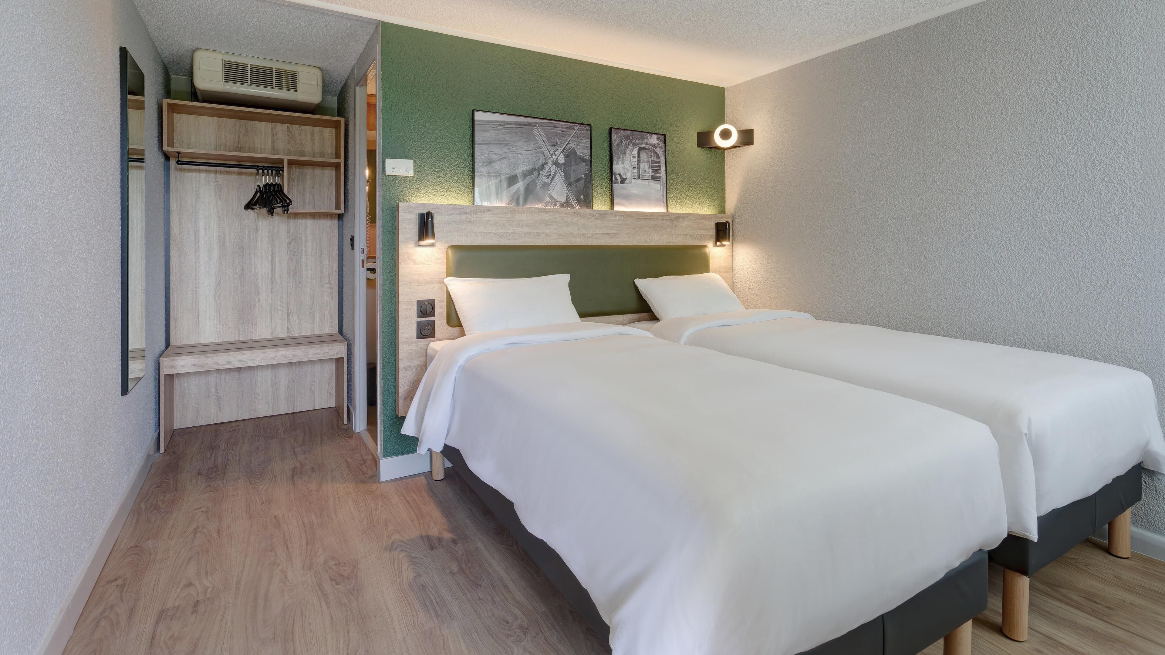 Een hotelkamer met twee bedden, een houten bankje, een kapstok en een wandairconditioner.