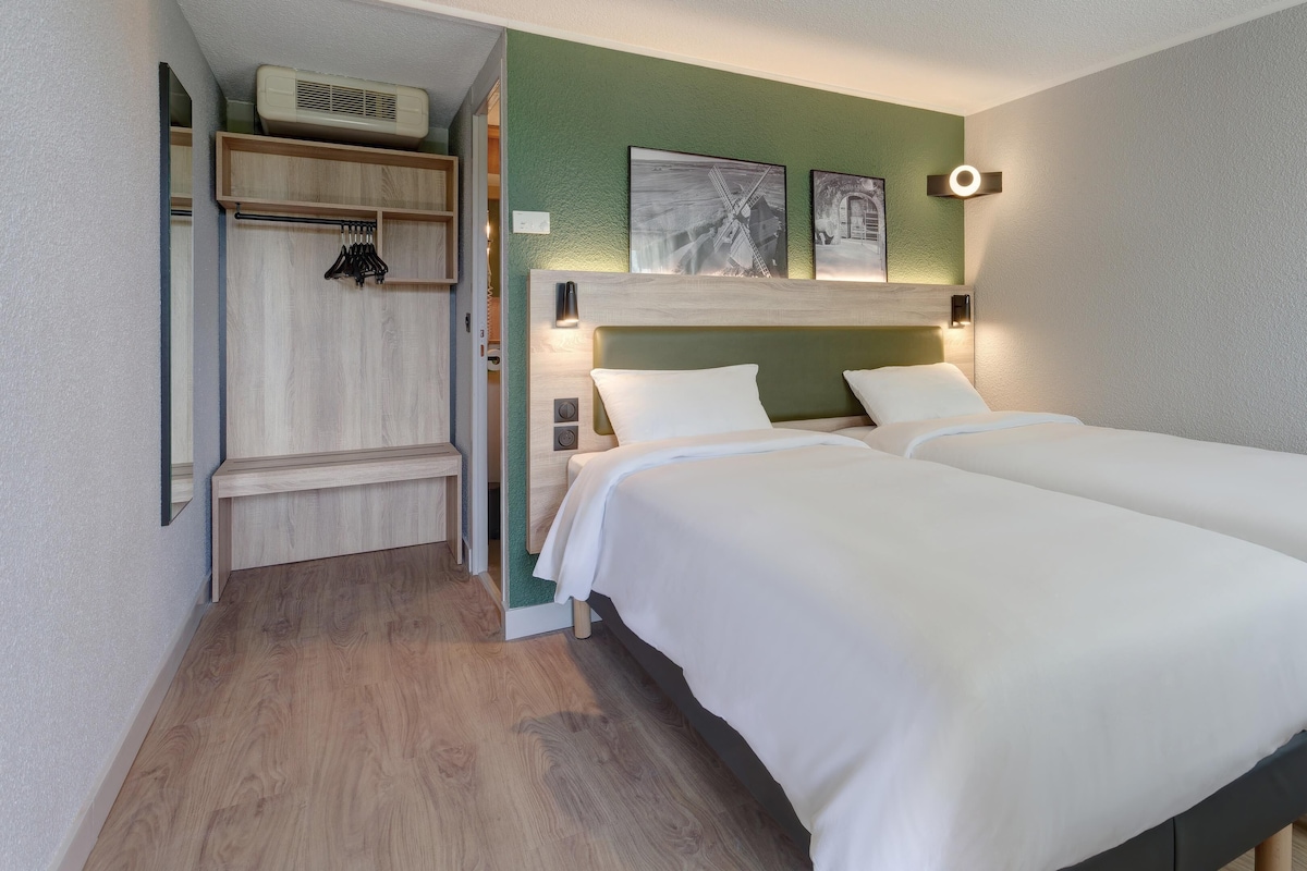 Habitación de hotel con dos camas, un banco de madera, perchero y aire acondicionado instalado en la pared.