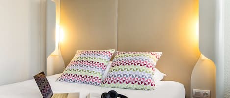 A neatly made bed with patterned pillows, a laptop, headphones, and a book on a white surface.