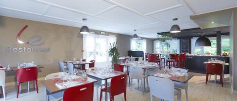 A restaurant with tables set for dining, red and white chairs, and a kitchen area visible in the background.