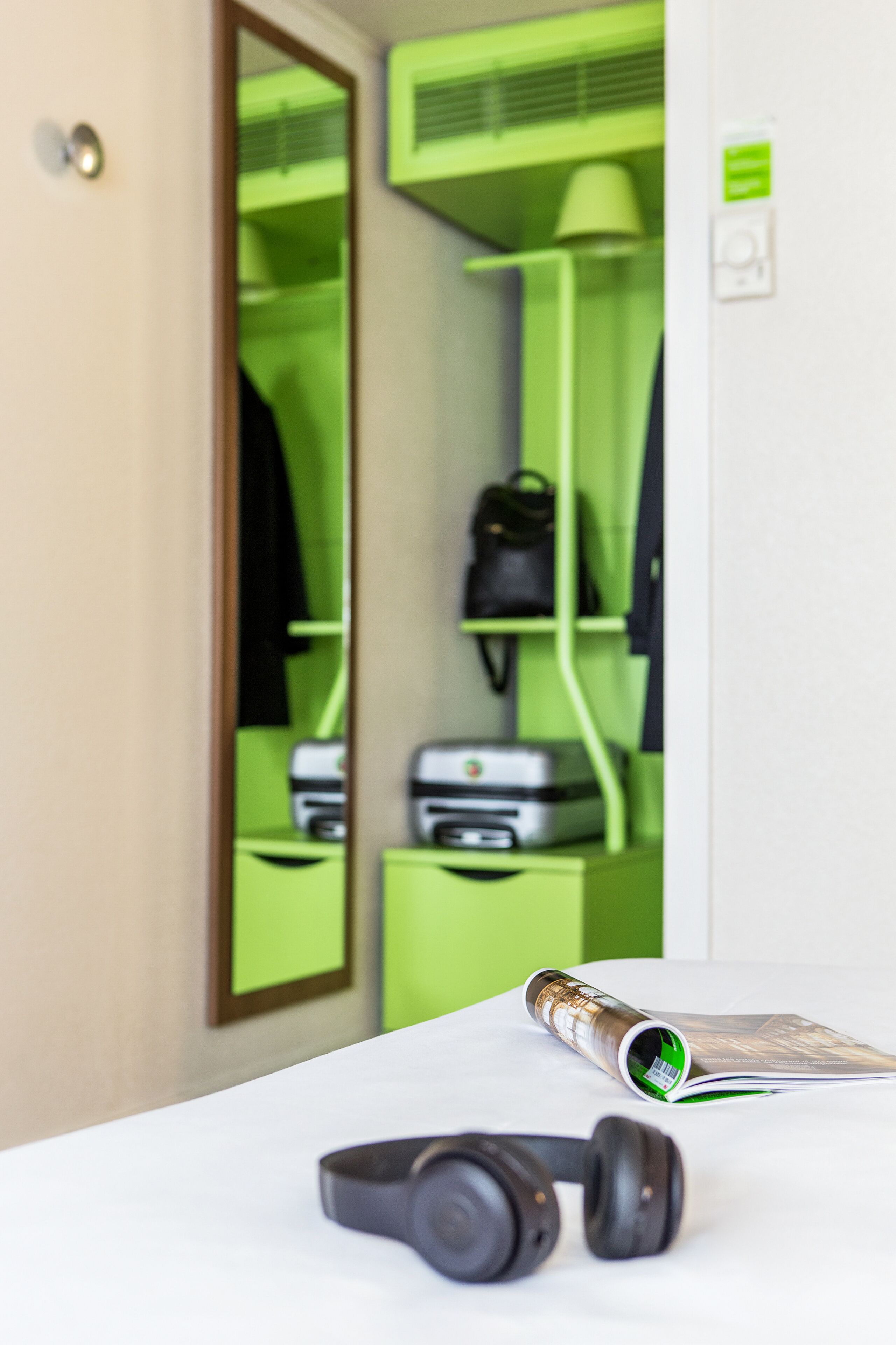 A room with a white desk, a pair of headphones, a rolled-up magazine, and a green wall with a mirror reflecting a green shelf with a black bag and a printer.