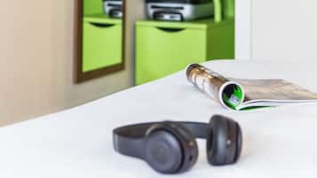 A room with a white desk, a pair of headphones, a rolled-up magazine, and a green wall with a mirror reflecting a green shelf with a black bag and a printer.