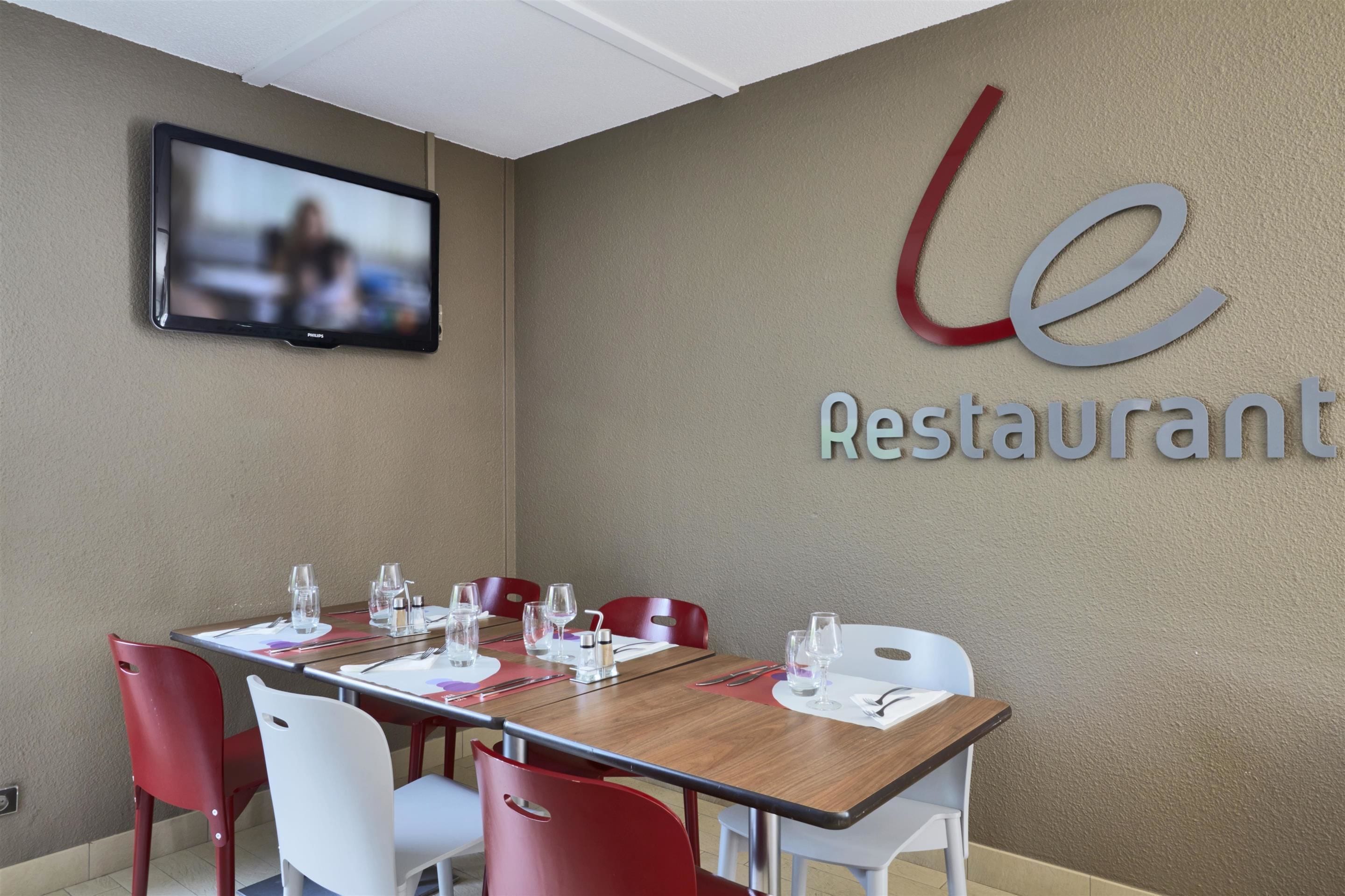 A restaurant with a long table set for dining, red and white chairs, and a mounted television on the wall.