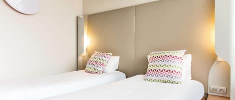 A hotel room with two beds, each with a patterned pillow, a bedside table, and a wall-mounted light fixture.
