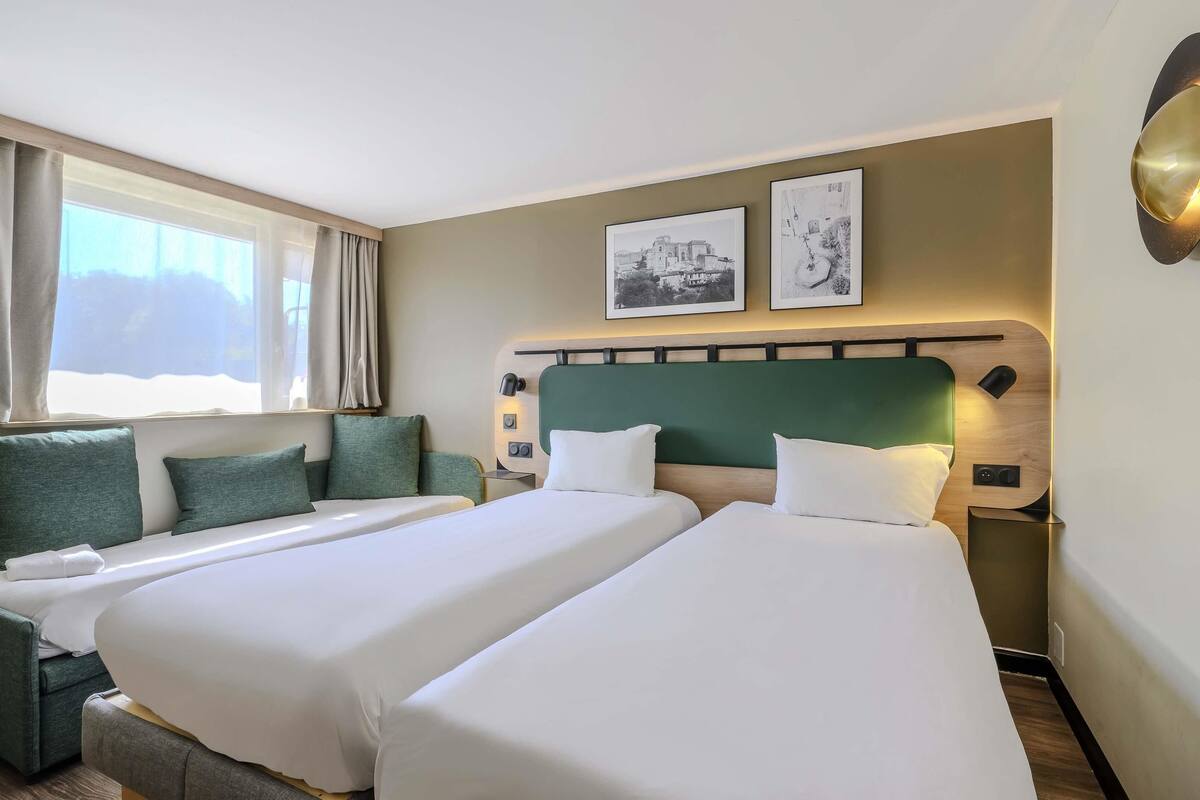 Habitación de hotel con dos camas, un sofá, una ventana con cortinas y dos cuadros en la pared.