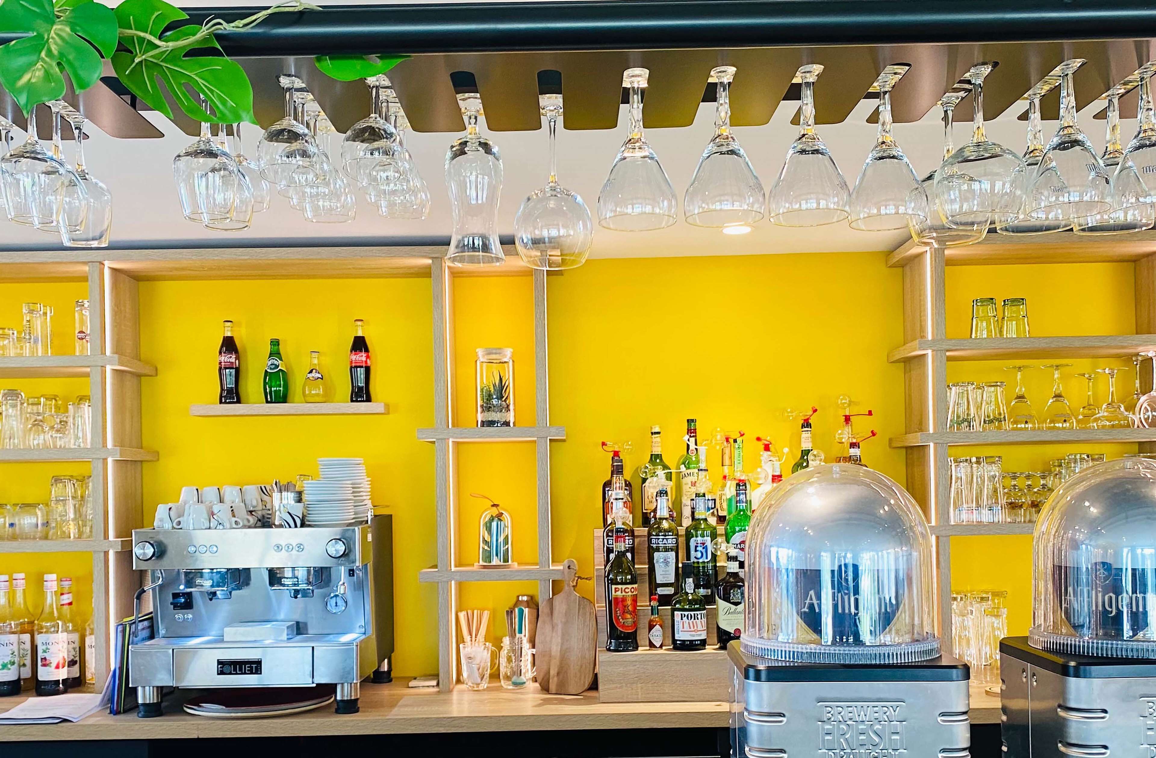Uma área de bar com parede amarela, prateleiras com garrafas, copos e uma máquina de café.