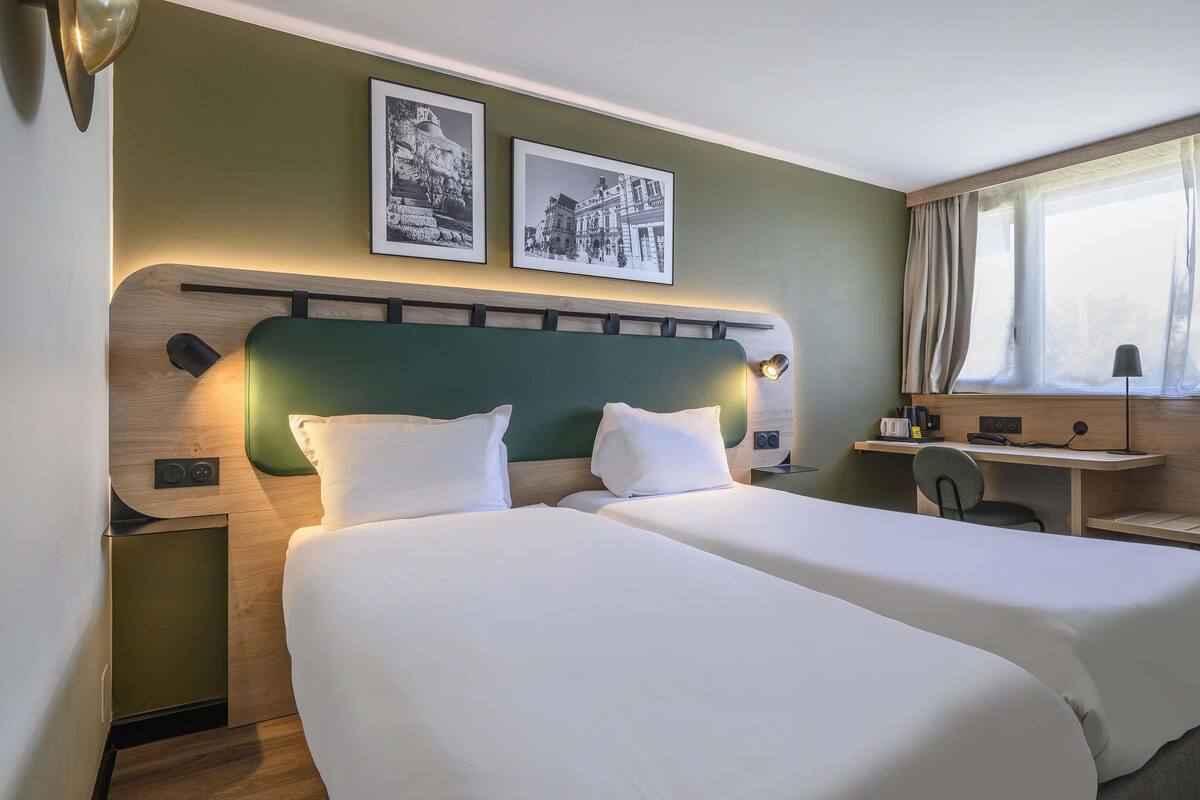 Habitación de hotel con dos camas, un escritorio, una silla, una ventana con cortinas y dos cuadros en la pared.