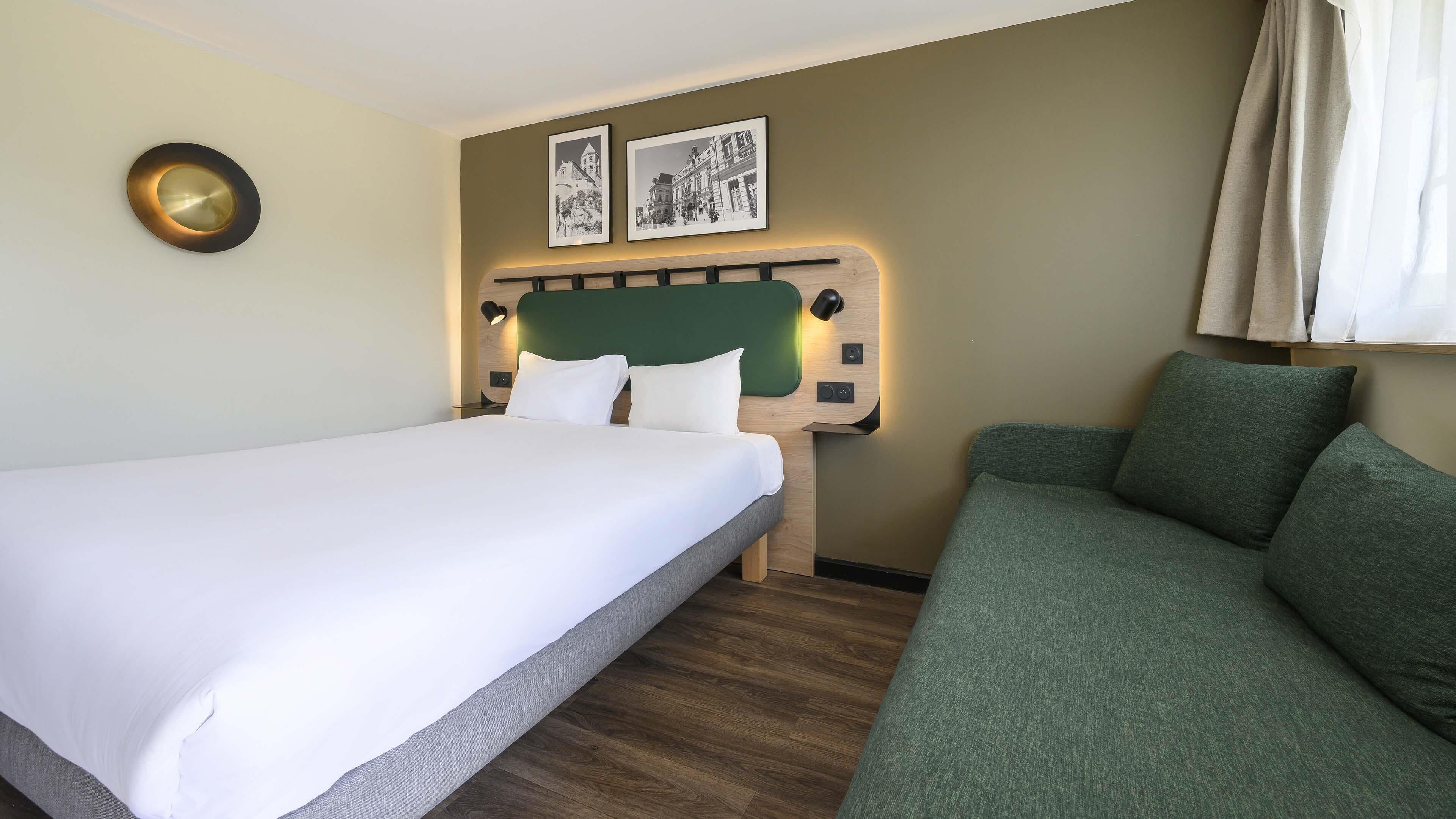 A hotel room with a bed, a green sofa, a headboard with wall art, and a wall-mounted clock.