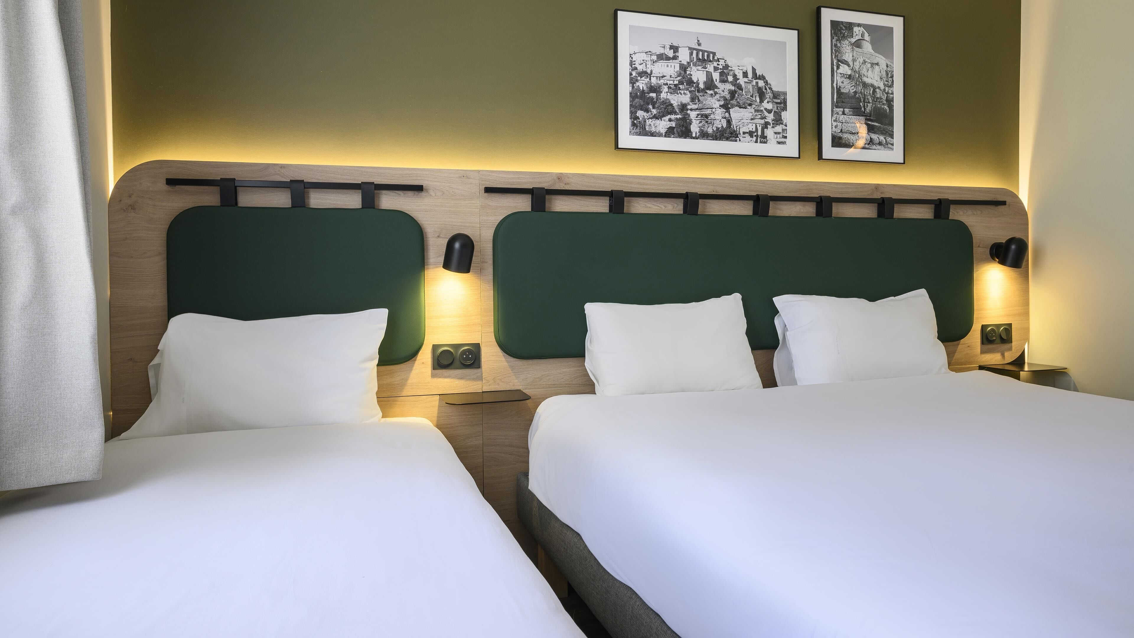 Ein Hotelzimmer mit zwei Betten, einem Holz-Kopfteil, Nachttischlampen und zwei schwarz-weißen Fotografien an der Wand.