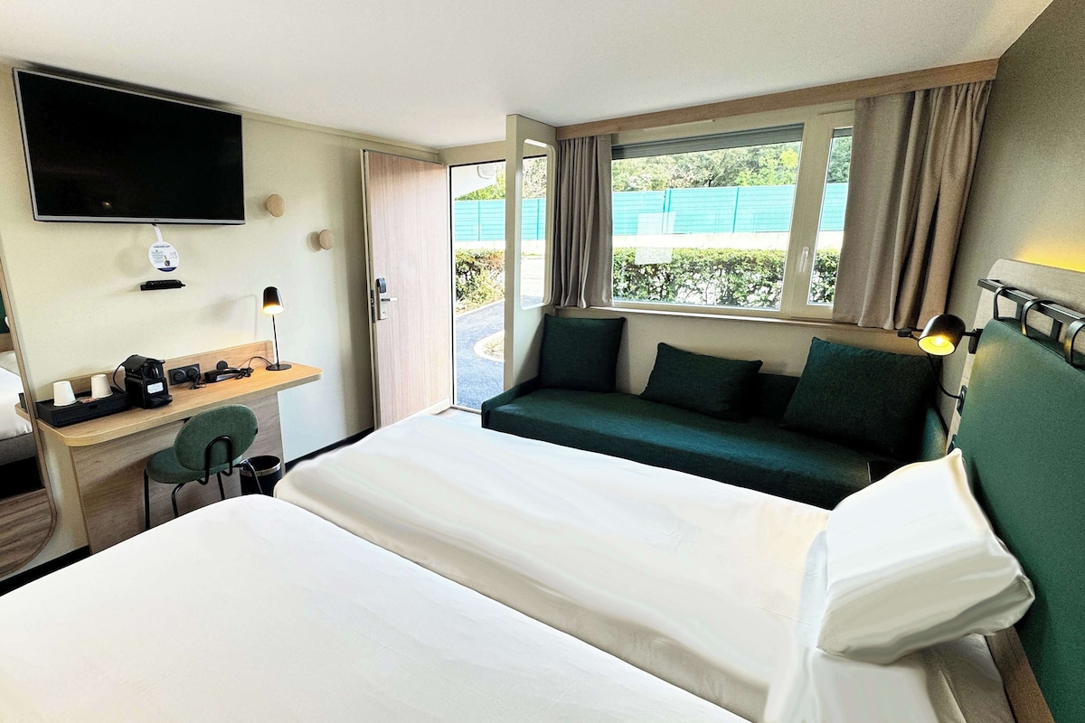 Habitación de hotel con una cama grande, un escritorio con silla, un televisor montado en la pared y una ventana con cortinas.