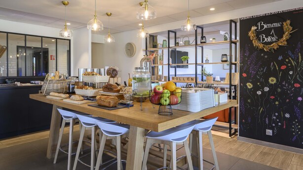 A café interior with a wooden counter, white bar stools, hanging pendant lights, and a wall with a floral mural. - Campanile NATURE - Bollène (Bollene)