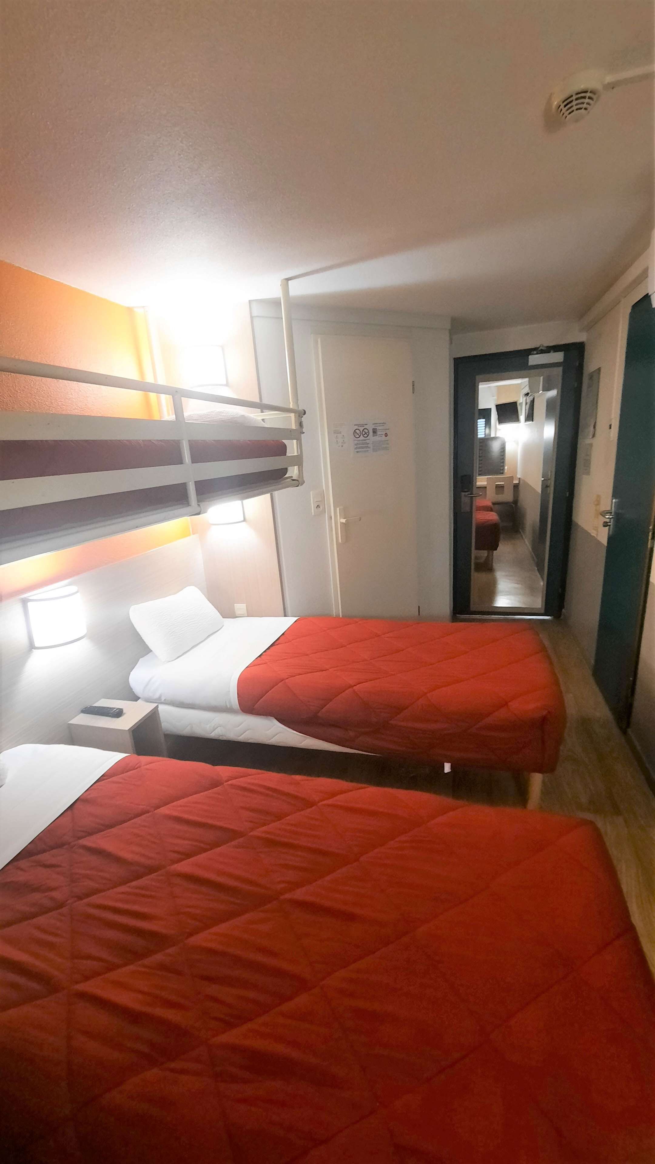 Habitación de hotel con dos camas individuales, una litera, una mesita de noche con lámpara y una puerta que da a un baño.