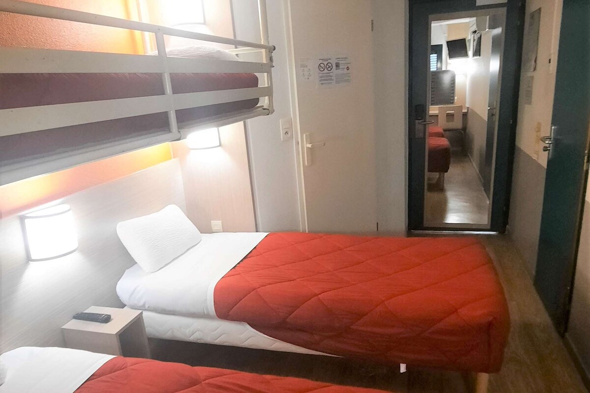 Habitación de hotel con dos camas individuales, una litera, una mesita de noche con lámpara y una puerta que da a un baño.