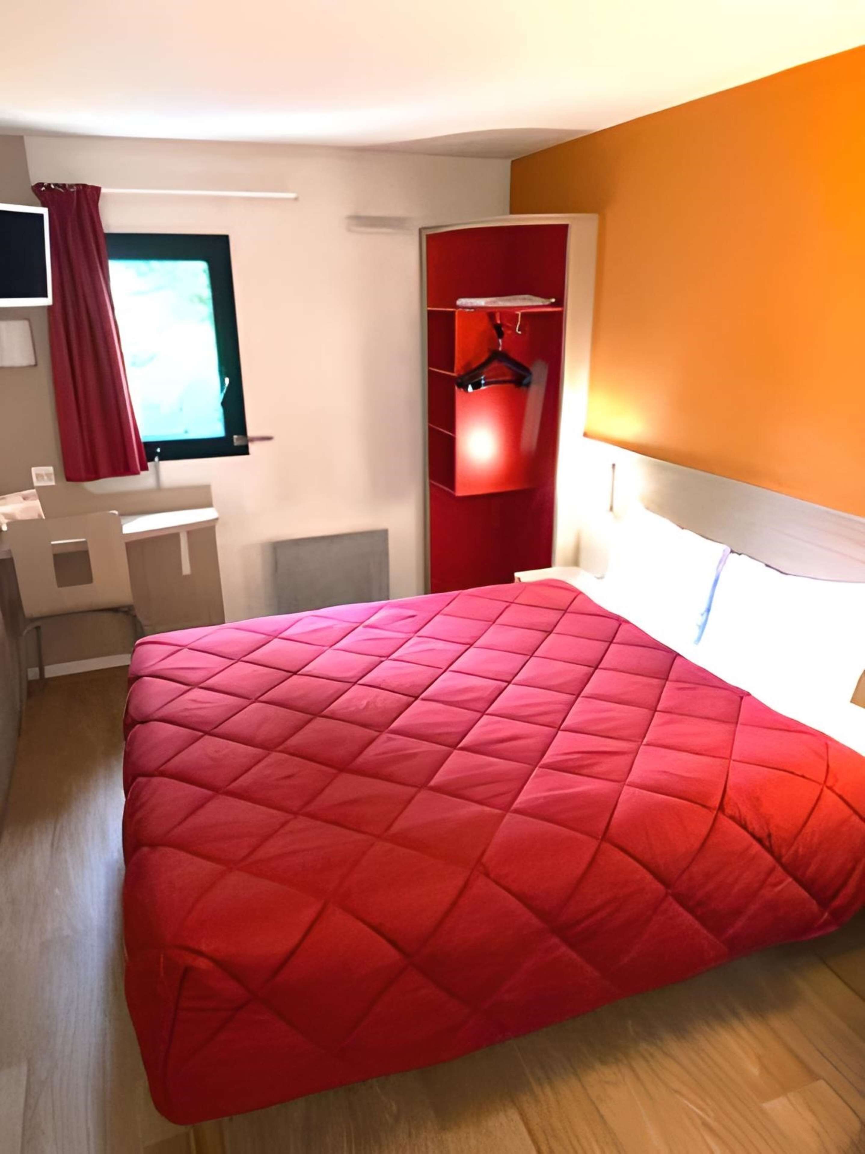 A modern bedroom with a red quilted bedspread, a desk, a chair, a TV, and a wardrobe with a red interior.