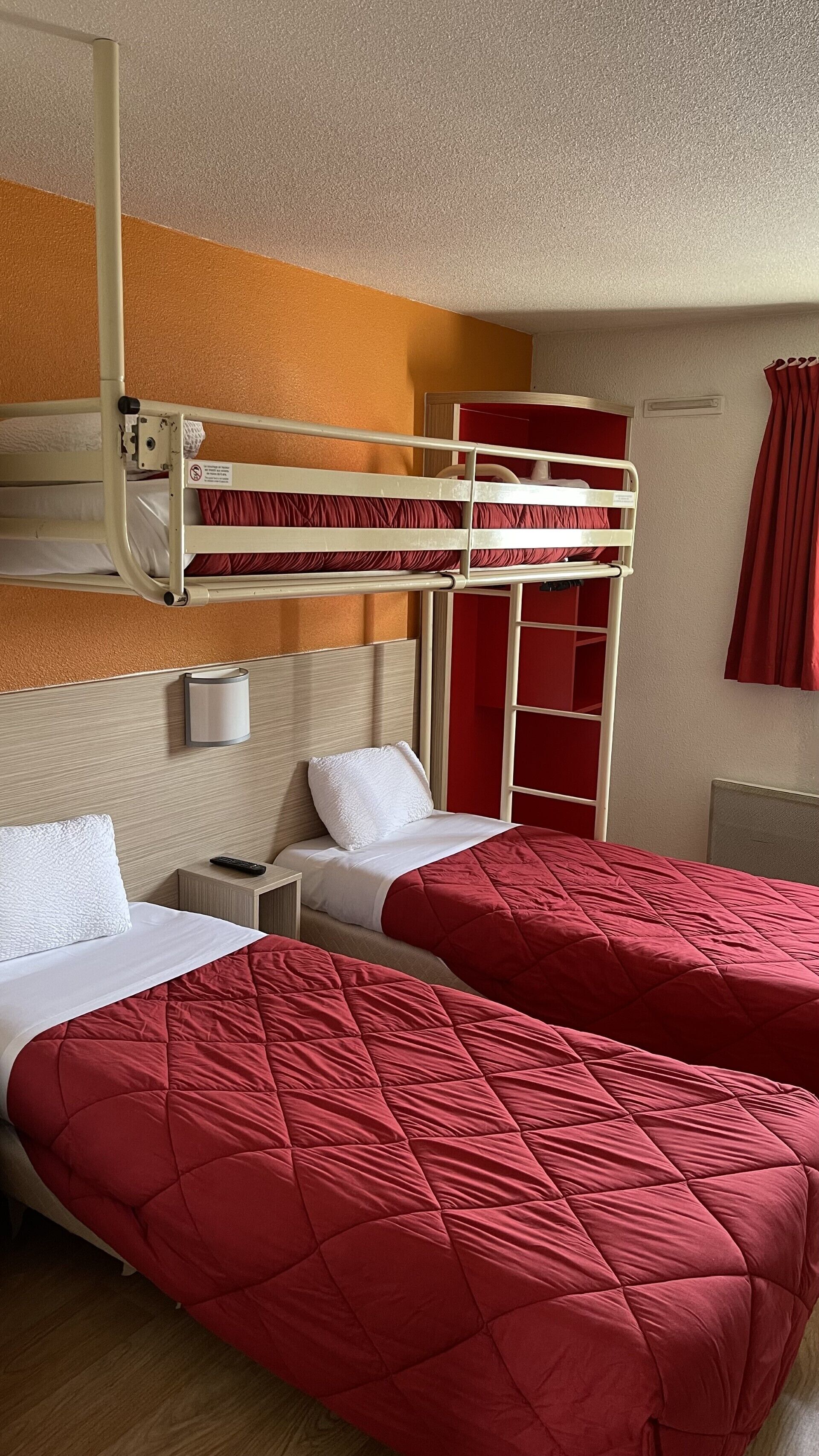 Habitación de hotel con literas, dos camas individuales, una mesita de noche, una lámpara de pared y una cortina roja.