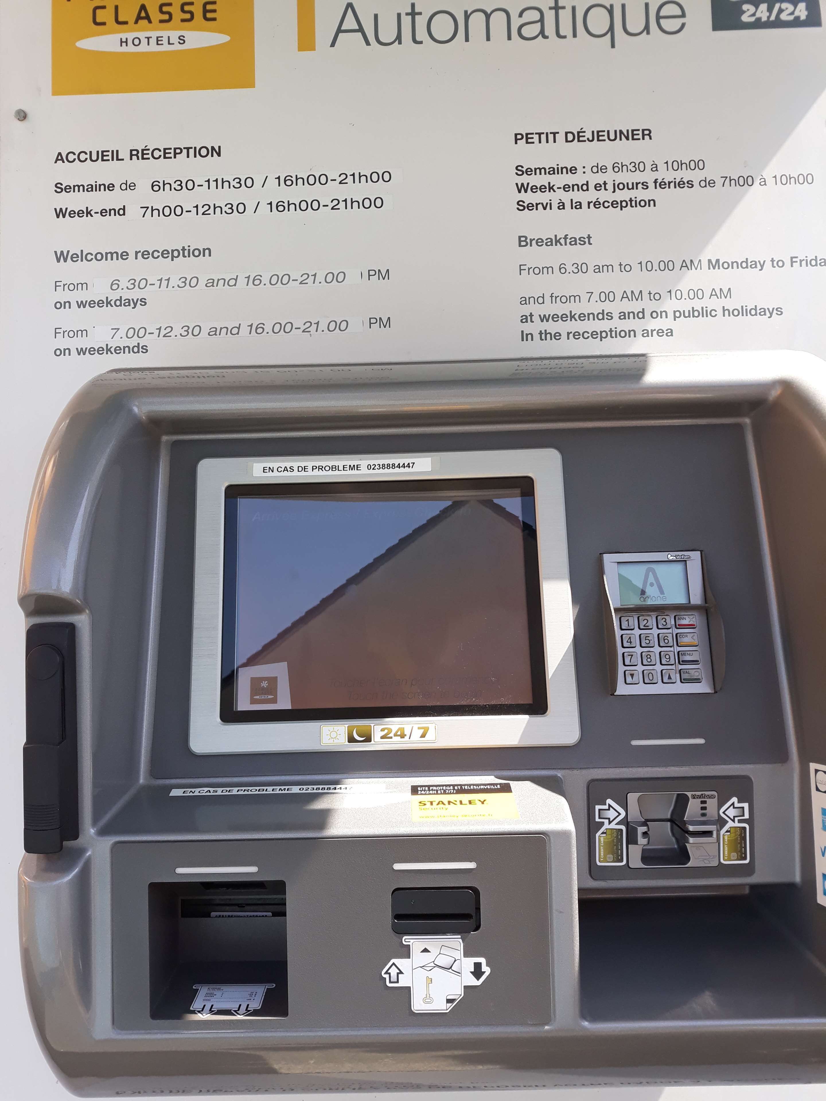 Een hotel receptiebalie met een digitaal scherm, een keypad en een bordje met 24/7 service.