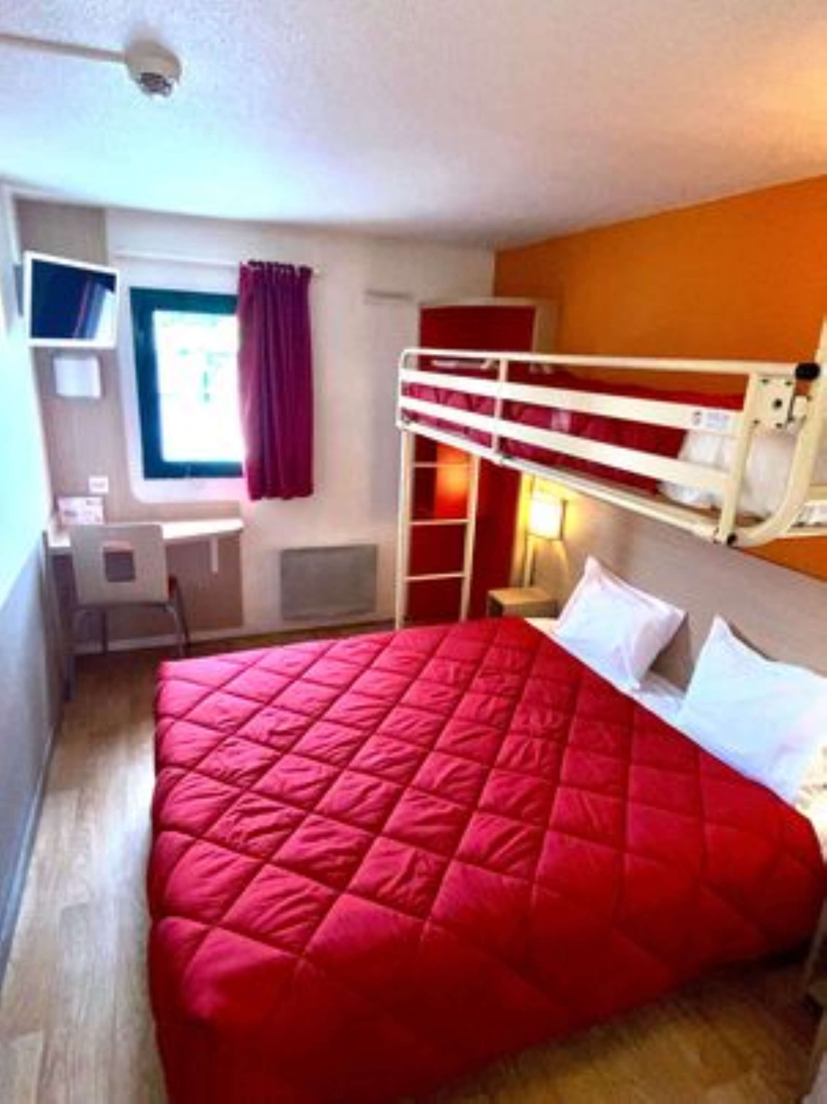 Una habitación con una cama litera, un escritorio con silla, un ventanal con cortinas y una cama con colcha roja.