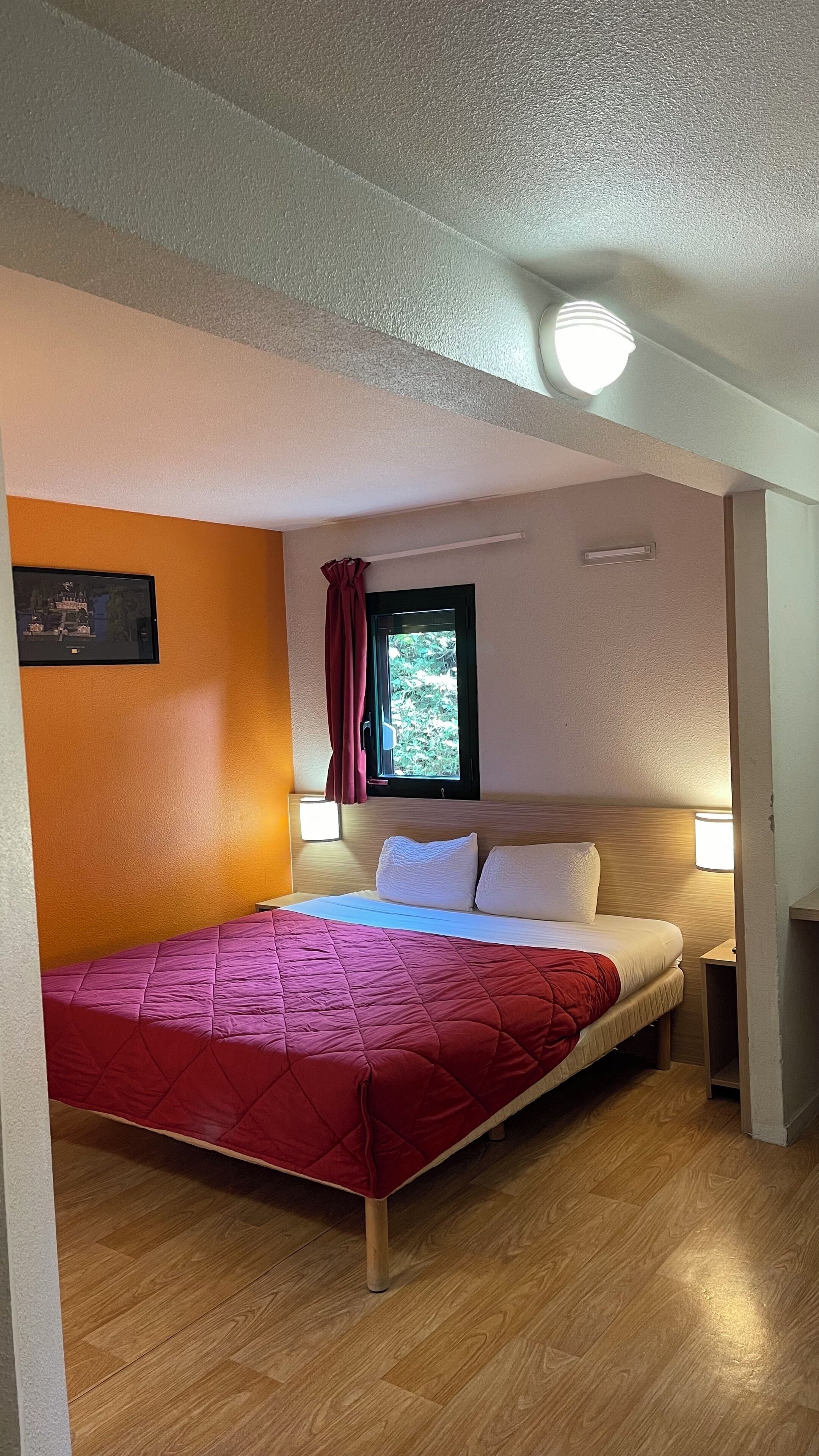 Un dormitorio con cama, cabecero de madera, ventana con vistas, televisor montado en la pared y una lámpara en el techo.