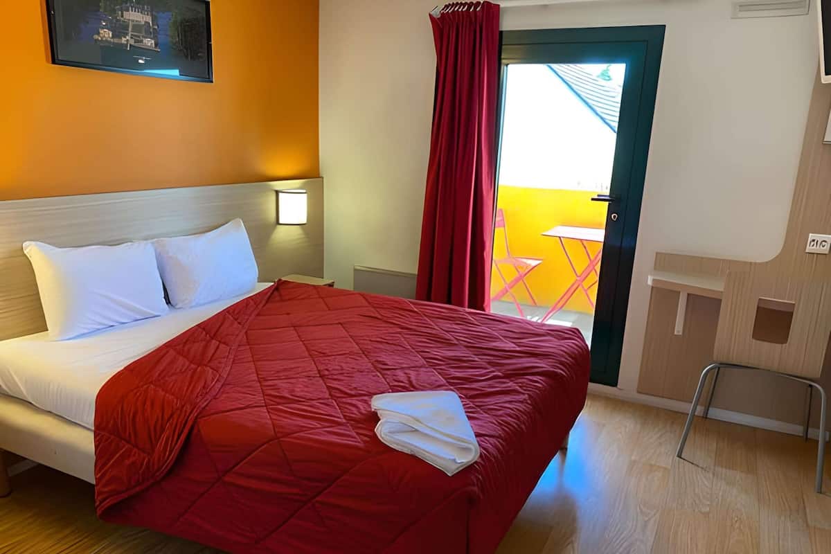 Habitación de hotel con cama, mesita de noche, televisor montado en la pared, ventana con cortina roja y vista a un edificio amarillo.
