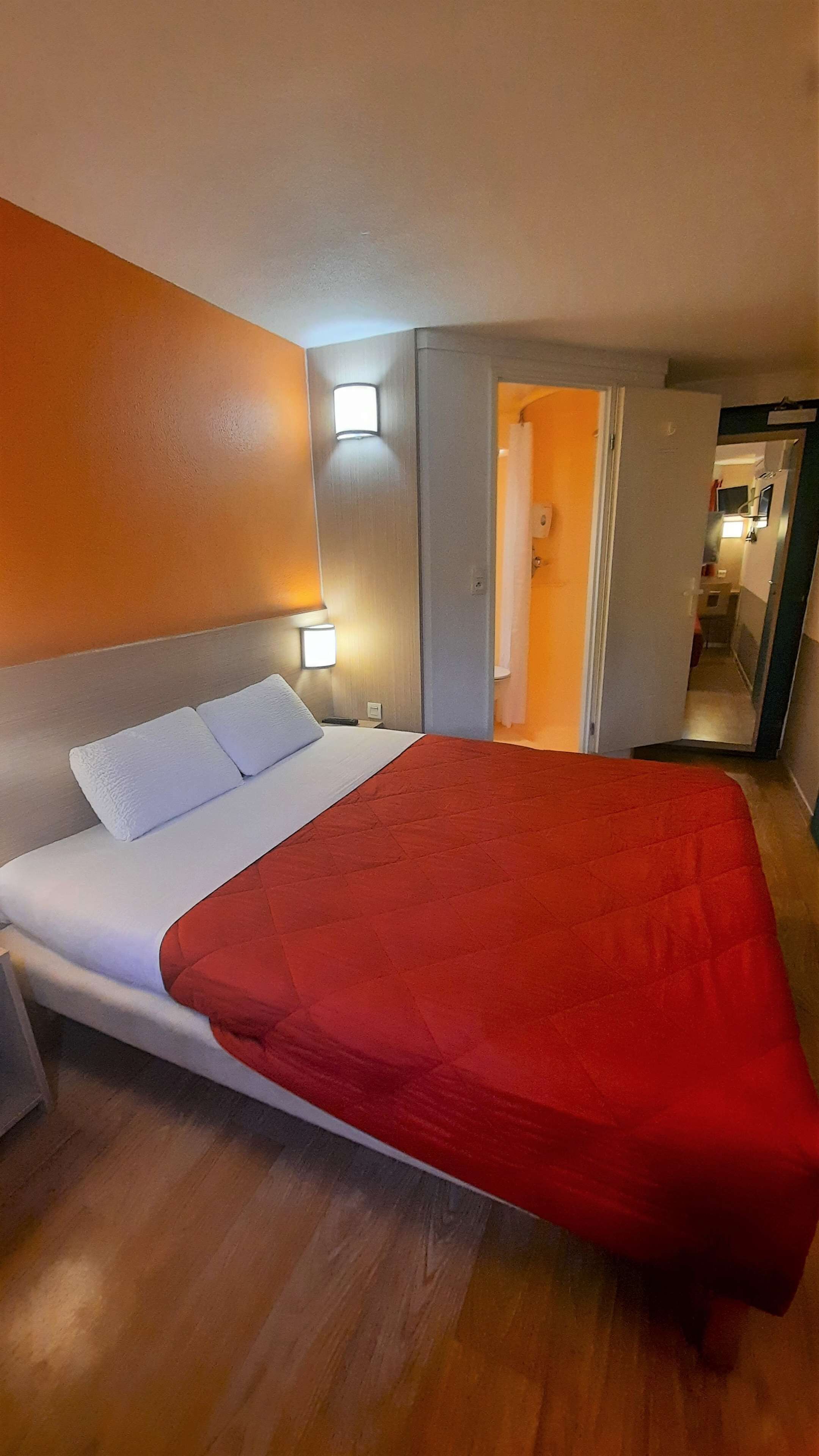A hotel room with a bed, a nightstand, a wall-mounted light, and an open door leading to a bathroom.