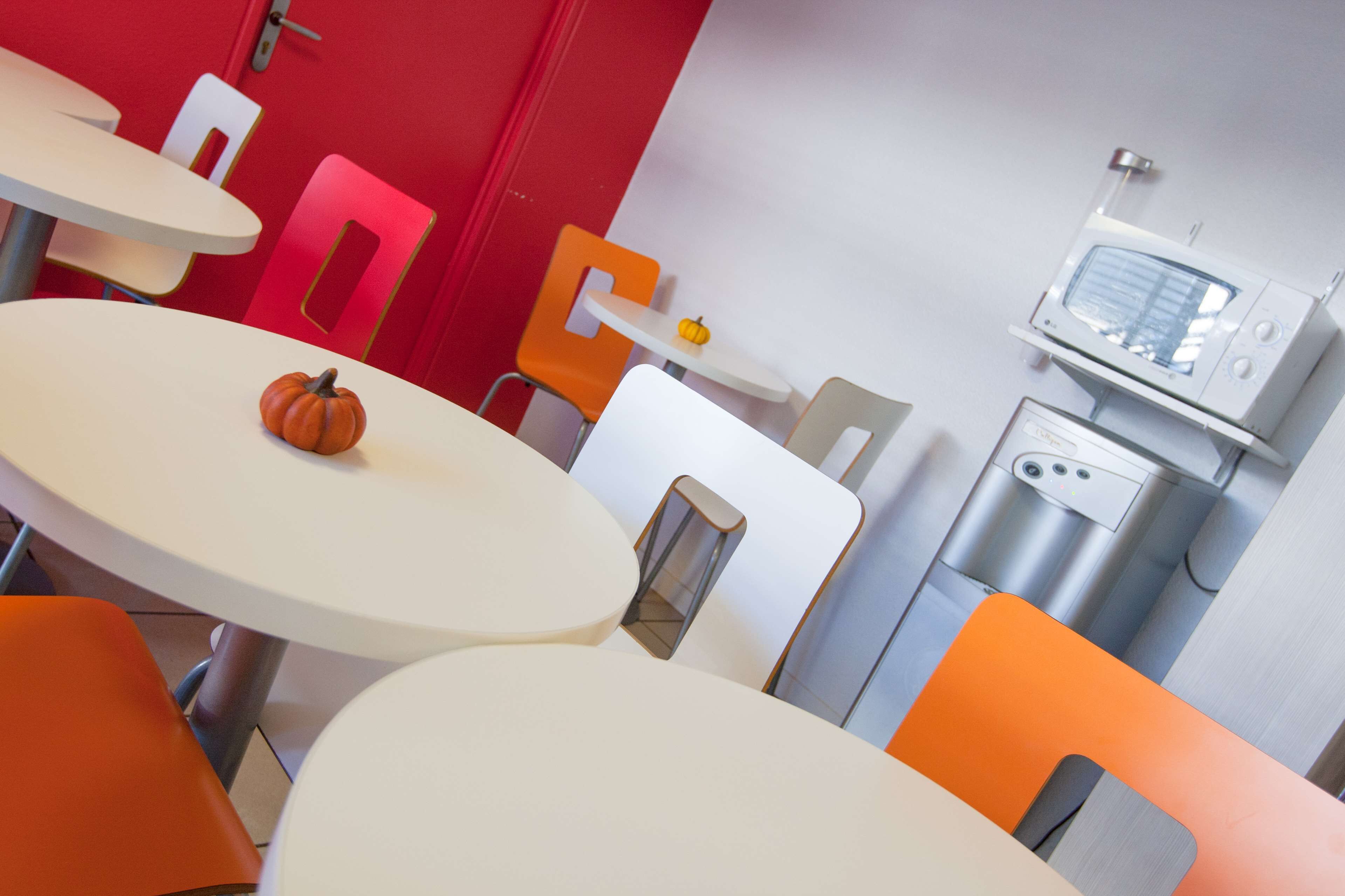 Un comedor moderno con mesas y sillas blancas y anaranjadas, un microondas y una pequeña decoración de calabaza.