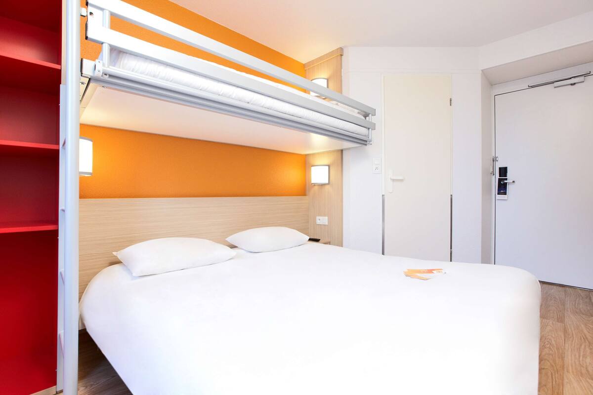 Habitación de hotel con una cama, un armario rojo, una puerta blanca y una lámpara fijada a la pared.