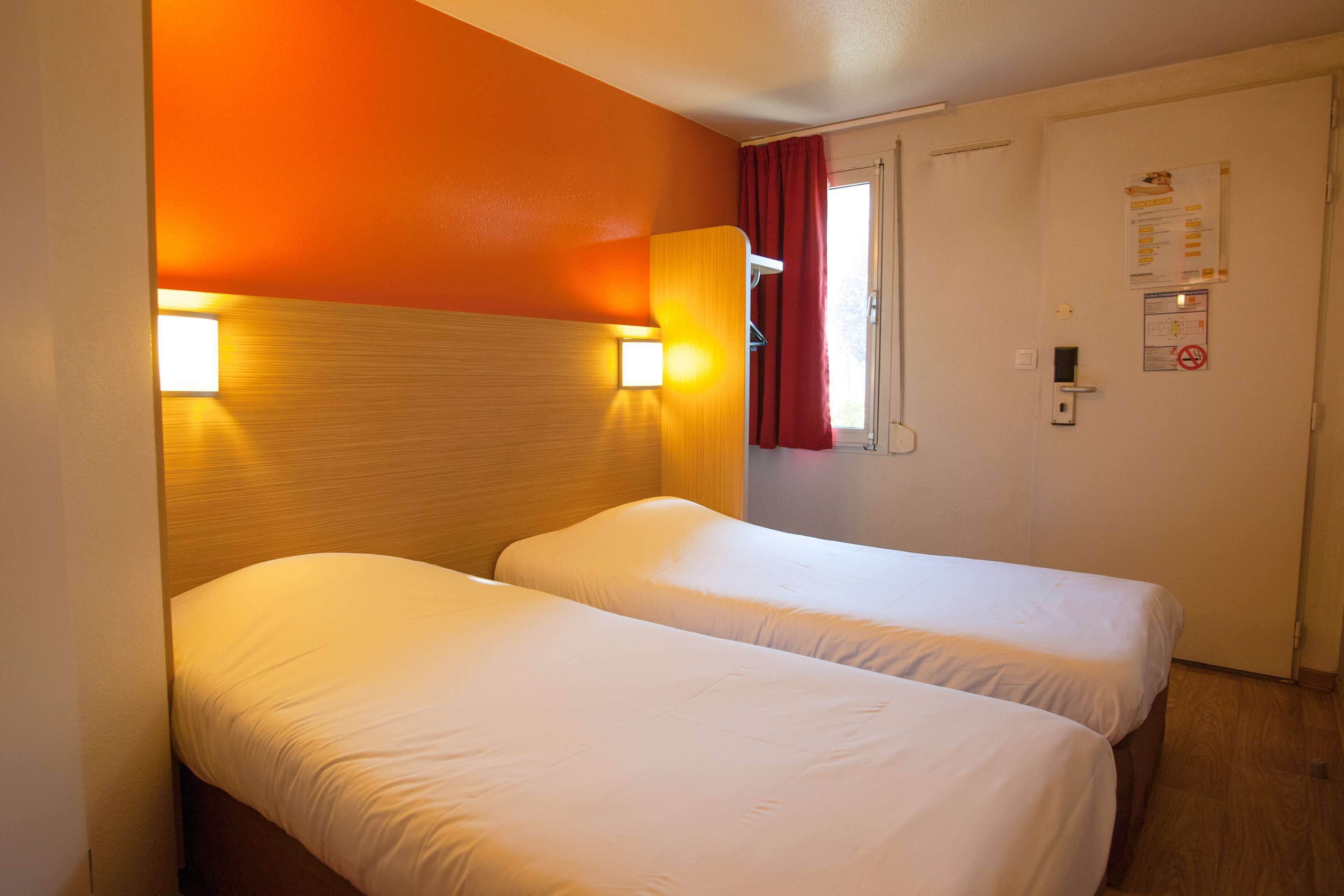 Een hotelkamer met twee enkele bedden, een houten hoofdbord, een wandlamp, een deur met een rood gordijn, en een bedieningspaneel aan de muur.