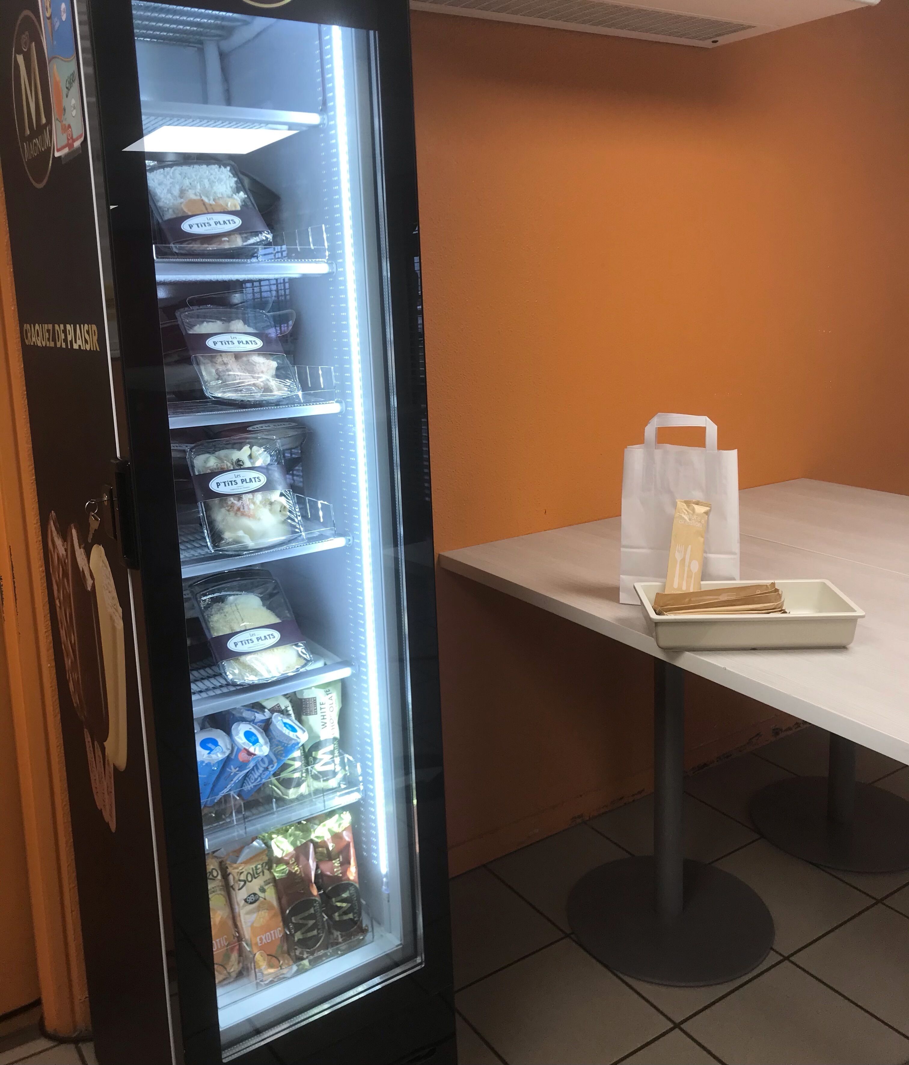 Un espositore self-service con diversi prodotti confezionati e un tavolo con un vassoio e sacchetti di carta.