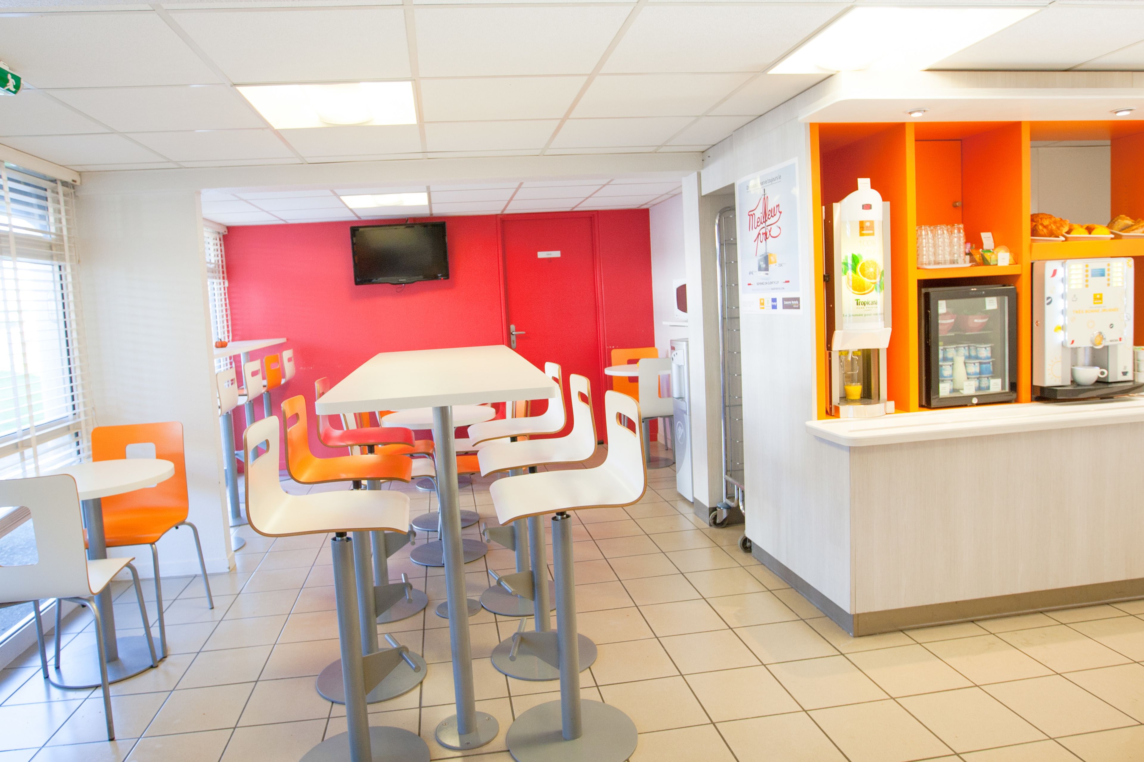 A brightly lit cafeteria with orange and white chairs, a white table, and a counter with a refrigerator and beverage dispenser.