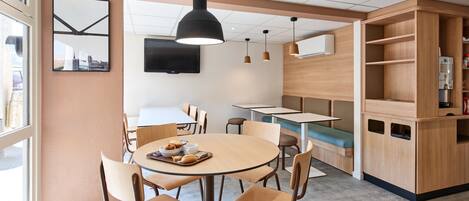 Un café moderno con mesas redondas, sillas de madera, una lámpara colgante grande y un televisor montado en la pared.