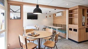 A modern cafe with round tables, wooden chairs, a large pendant light, and a wall-mounted TV.