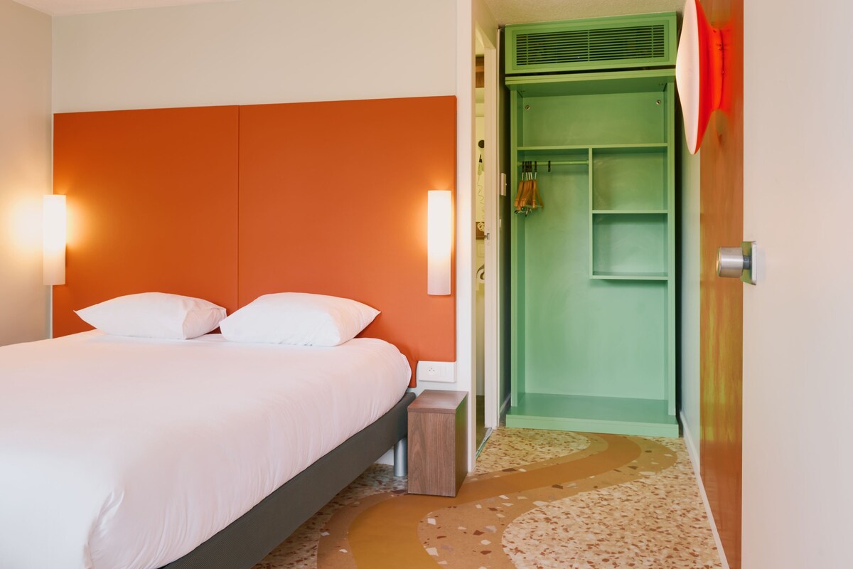 A hotel room with a bed, a green wardrobe, and a small trash bin.