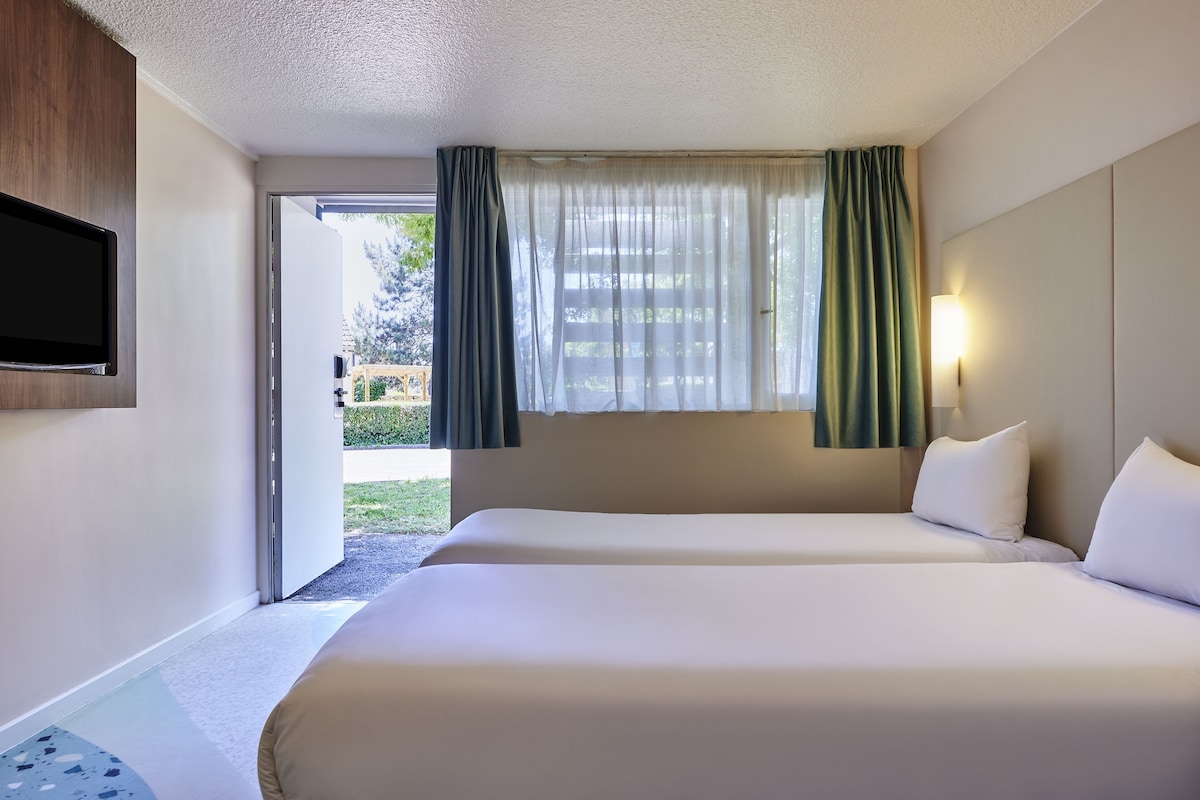 A hotel room with two beds, a TV mounted on the wall, a window with curtains, and a view of the outdoors.