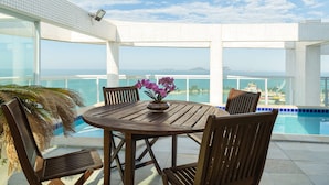 A wooden table with chairs and a vase of flowers on a balcony overlooking a pool and the ocean. - Golden Tulip Macaé (Macaé)
