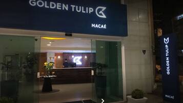 Exterior de un hotel de diseño moderno llamado 'Golden Tulip Macaé', con recepción visible a través de puertas de vidrio.