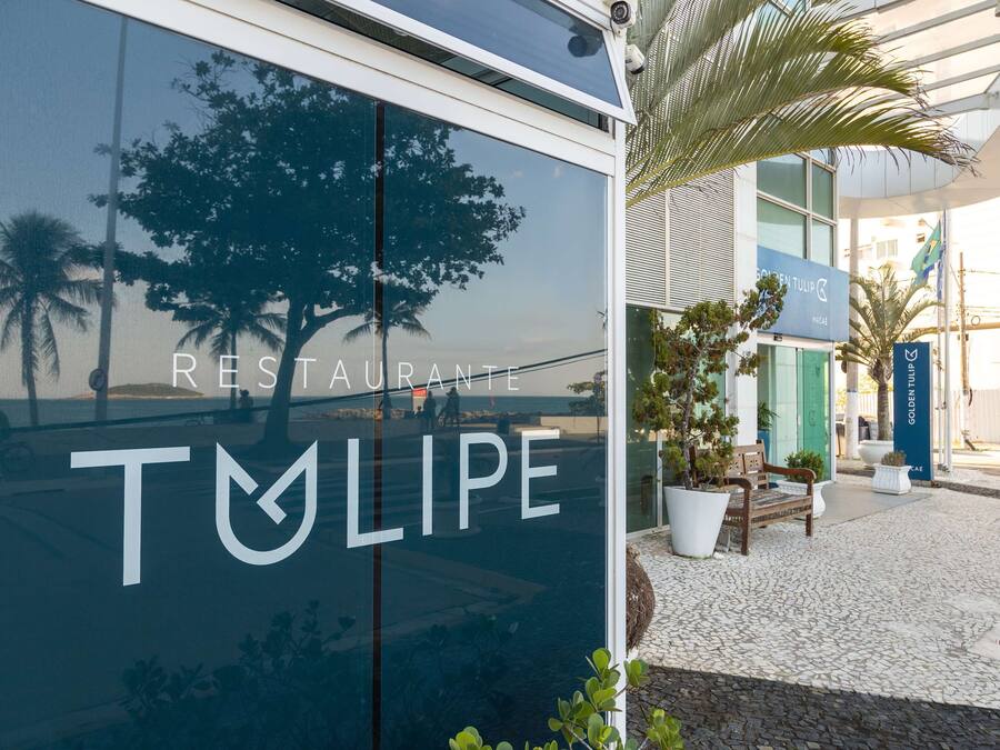 Um edifício moderno com uma placa indicando "RESTAURANTE TULIPE" e o reflexo de palmeiras em sua fachada de vidro.