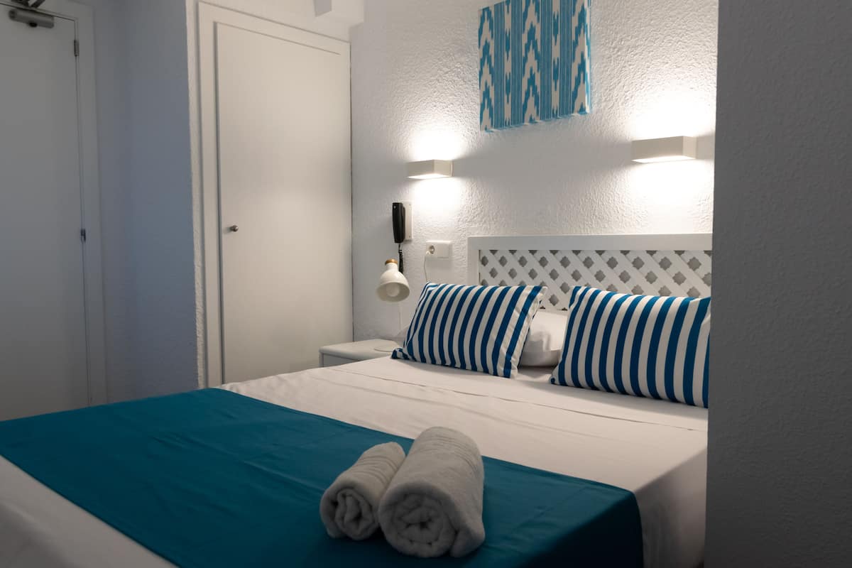 A neatly made bed with a teal bedspread and two rolled towels, a white headboard, and a wall-mounted light fixture.