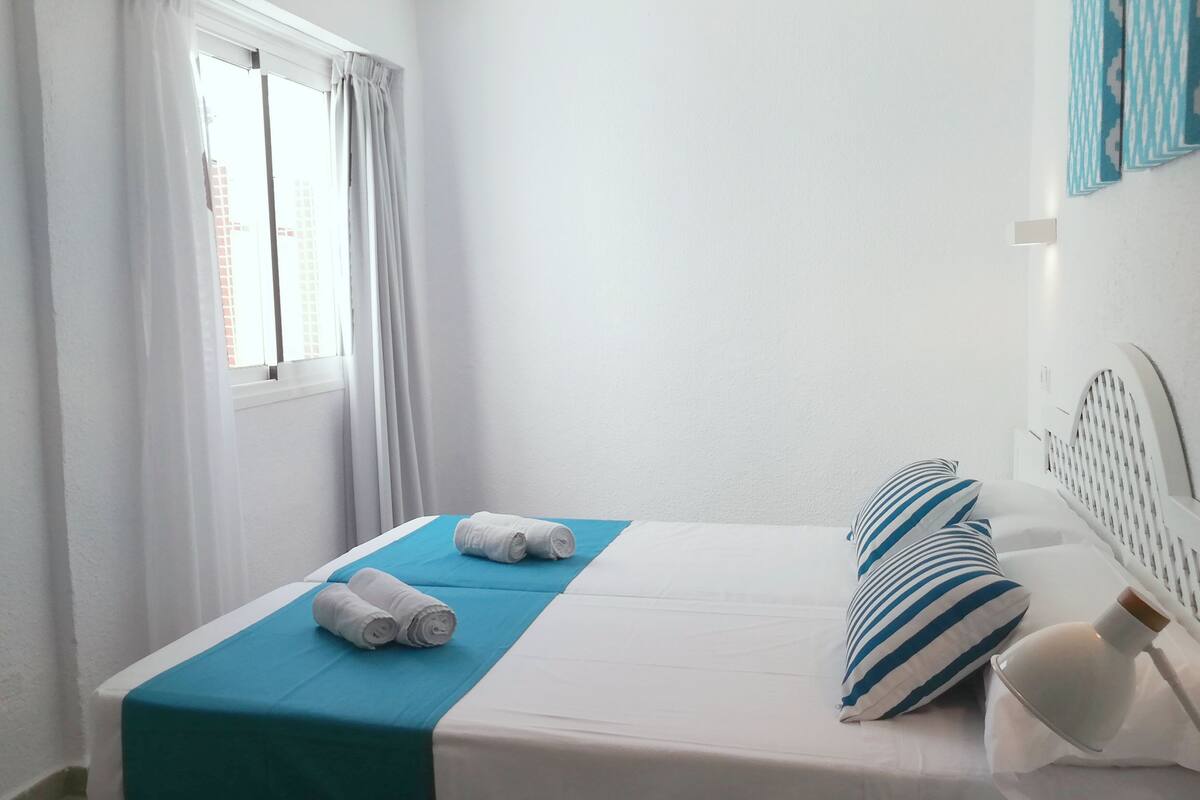 A neatly made bed with a blue bedspread and white pillows, a window with white curtains, and a white lamp on the bedside table.