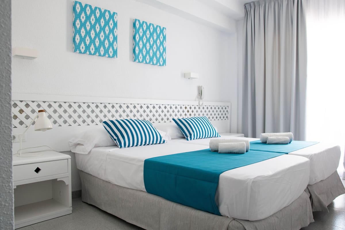 A double bed with a teal bedspread, two striped pillows, and a white nightstand with a lamp.