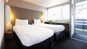 Una habitación de hotel con dos camas, un escritorio, una silla, una lámpara y un ventanal con cortinas.
