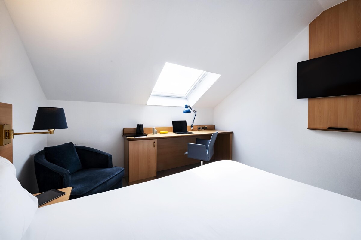 A modern hotel room with a skylight, a desk with a laptop, a chair, and a bed.