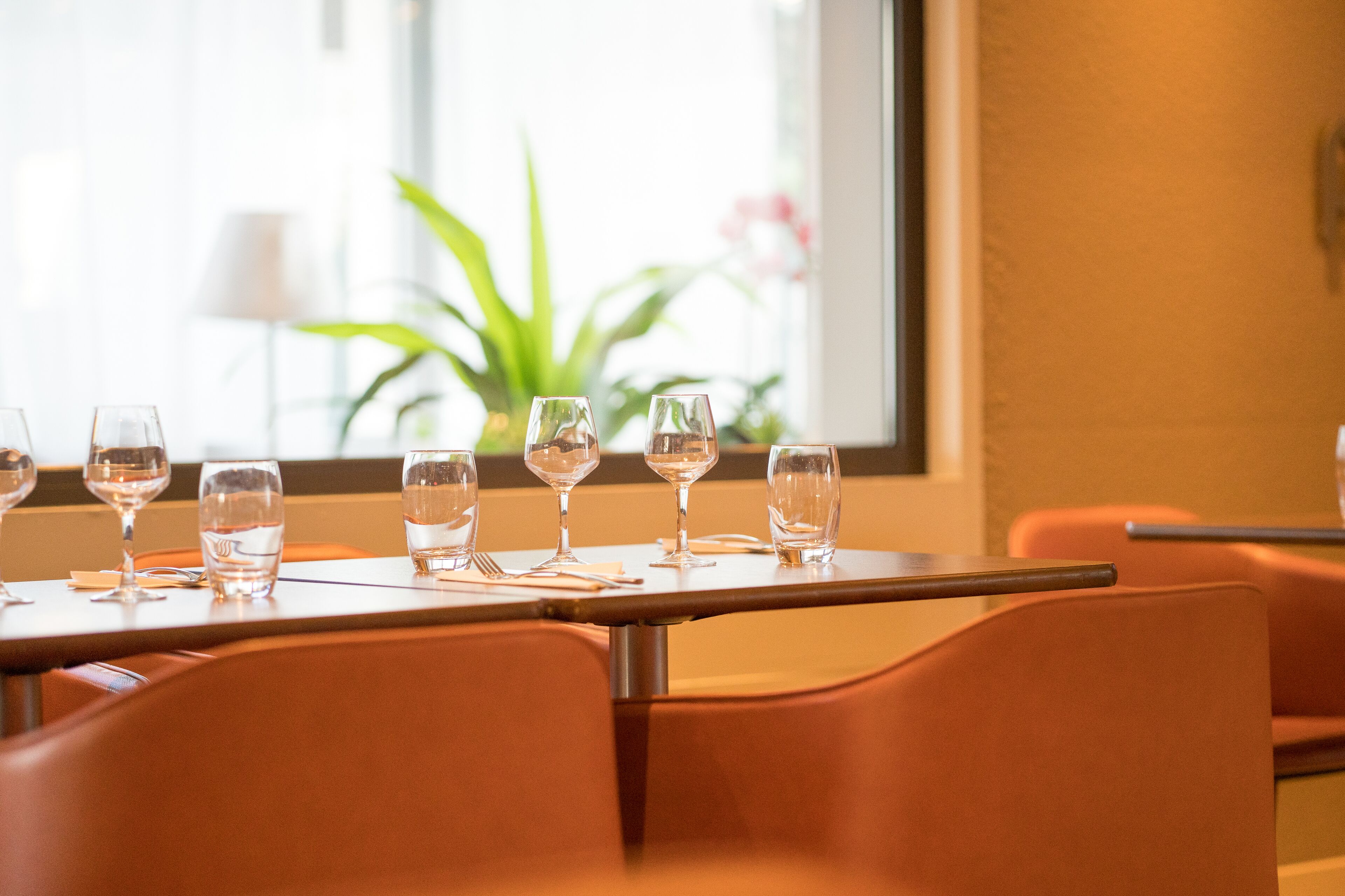Una mesa de comedor con seis copas de vino y cubiertos, ubicada en una habitación con sillas anaranjadas y una ventana con una planta al fondo.