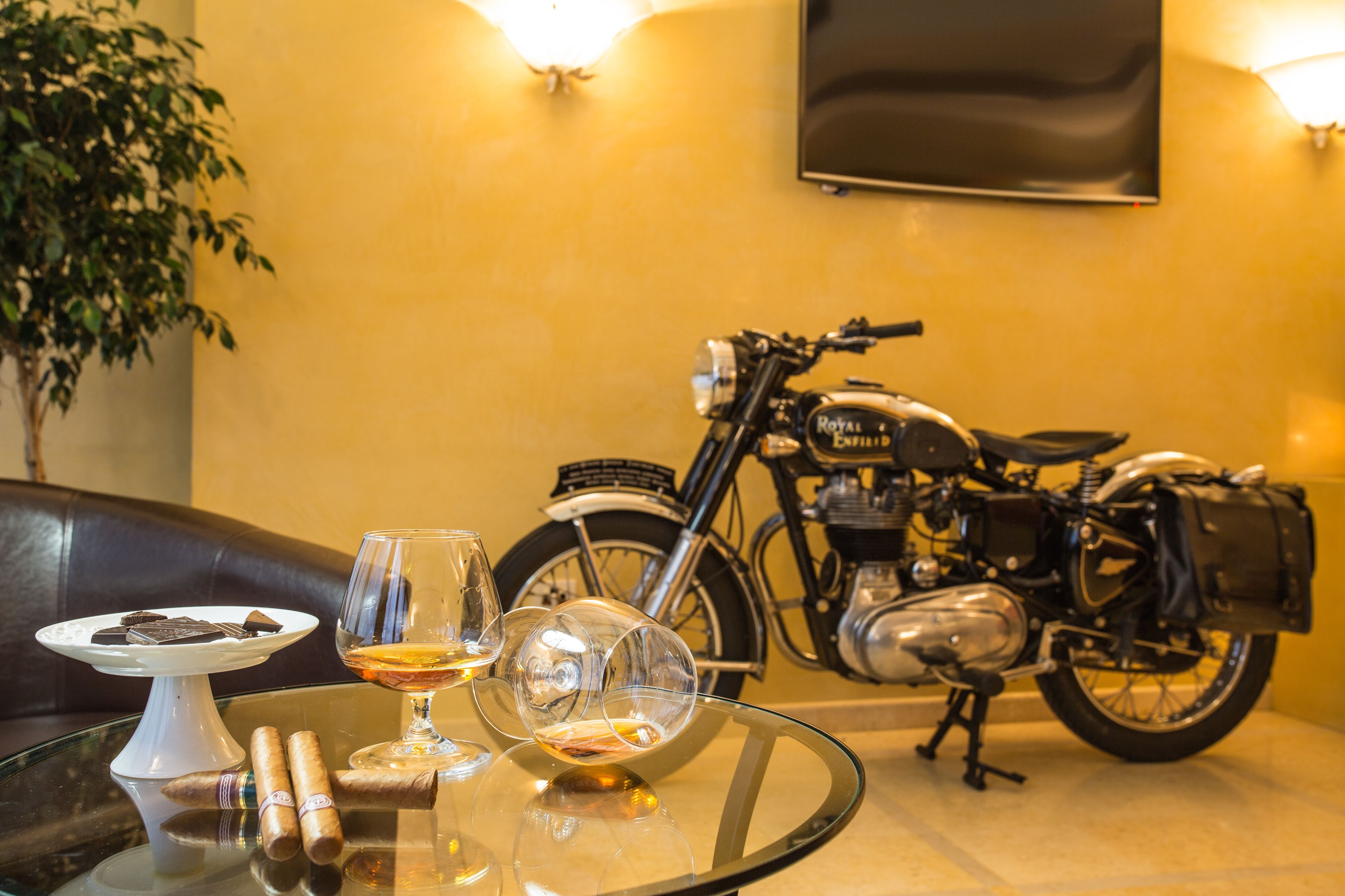 A glass table with a plate of chocolates, two glasses, and cigars. A vintage motorcycle is parked in the background.