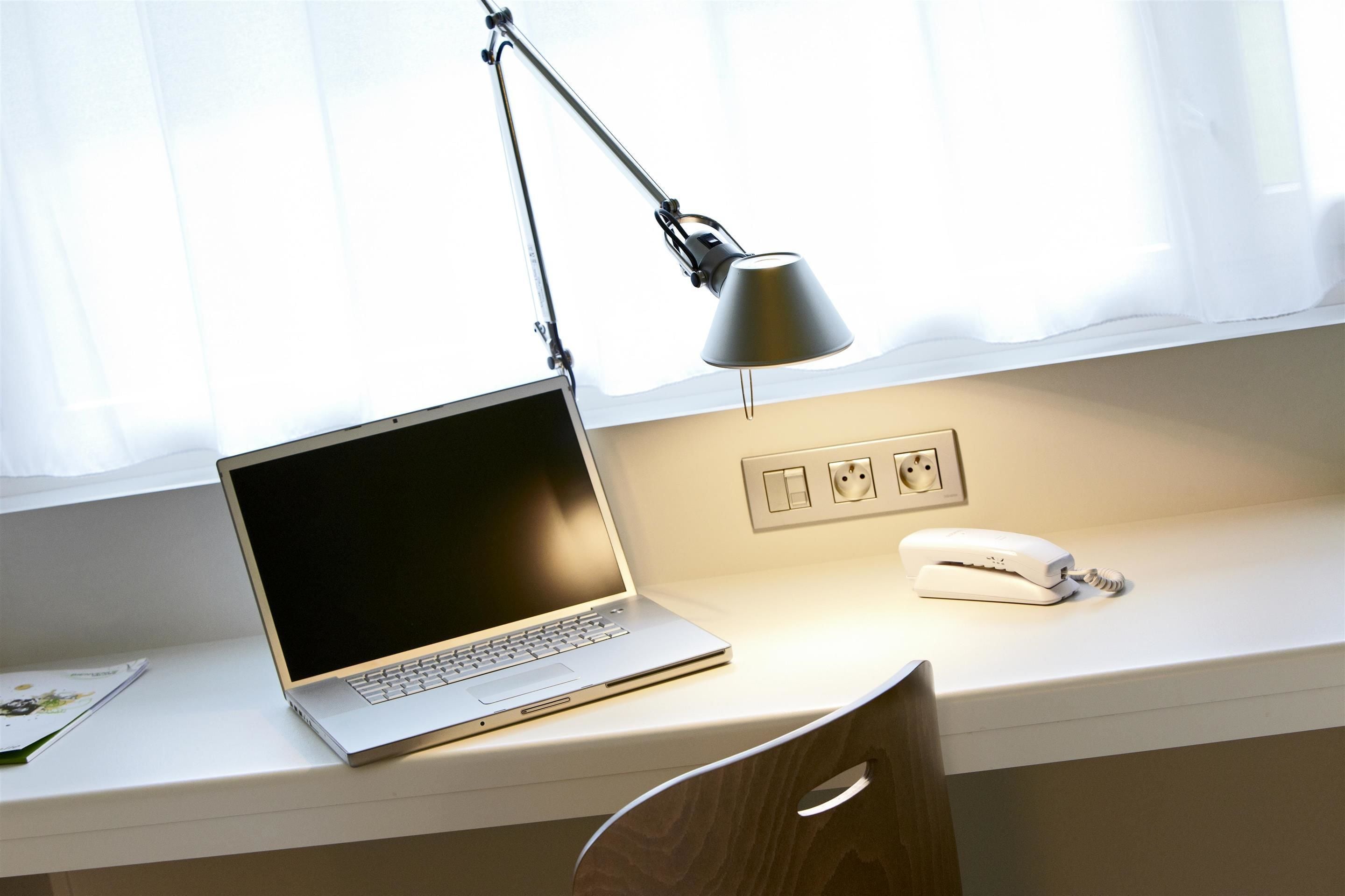 A laptop on a desk with a desk lamp, a phone, and a window with curtains.