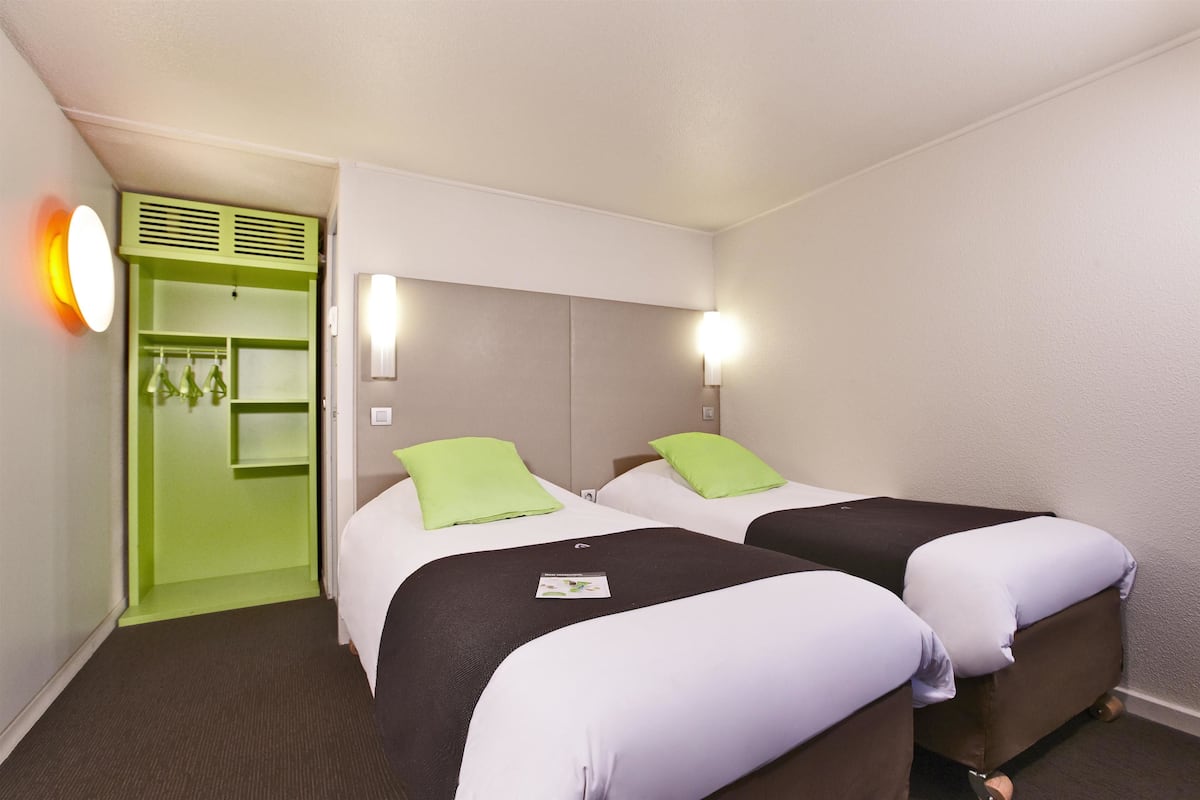 A hotel room with two beds, each with a pillow and a blanket, a green wardrobe, and a wall-mounted light fixture.