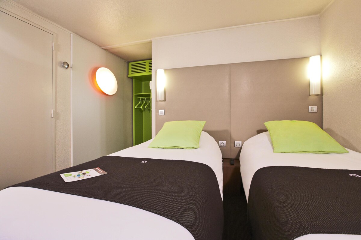 Une chambre d’hôtel avec deux lits, chacun étant équipé d’un oreiller et d’une couverture. On y trouve un placard avec un miroir et un luminaire au-dessus.