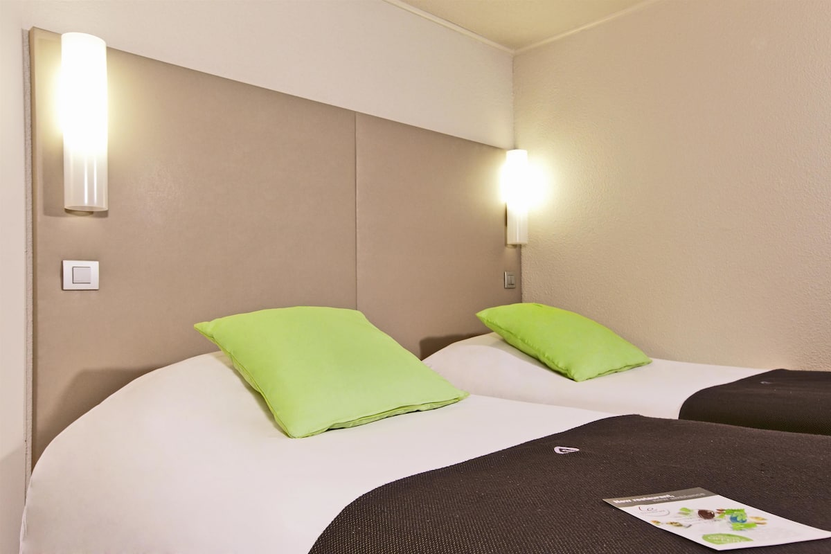 A hotel room with two beds, each with a green pillow, a black headboard, and a light fixture on the wall.