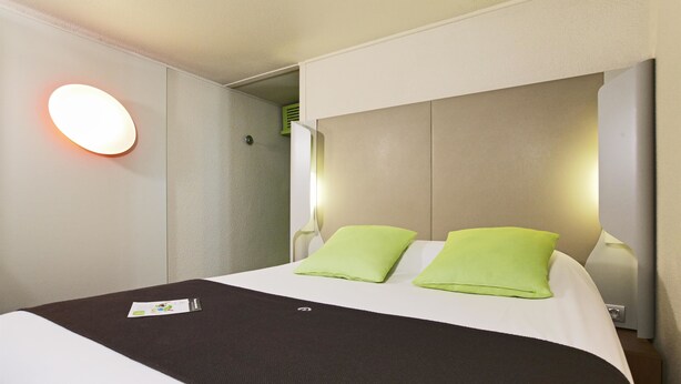 Une chambre d’hôtel avec un lit, deux oreillers verts, une tête de lit blanche et une lampe fixée au mur.