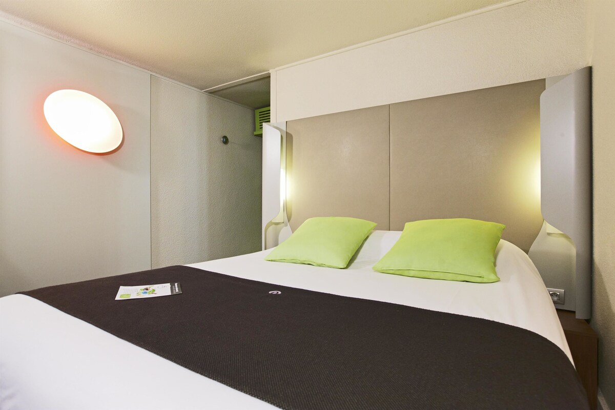 Une chambre d’hôtel avec un lit, deux oreillers verts, une tête de lit blanche et une lampe fixée au mur.